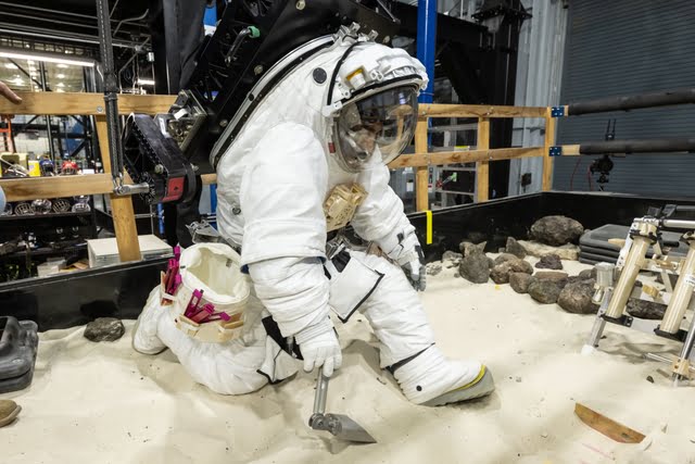 A NASA crew member practices using lunar tools to collect geology samples at NASA’s Johnson Space Center during an elevated suit pressure test where teams evaluate how well crew perform tasks in different suit pressure levels while wearing the Artemis III lunar spacesuit developed by Axiom Space called the AxEMU (Axiom Extravehicular Mobility Unit).